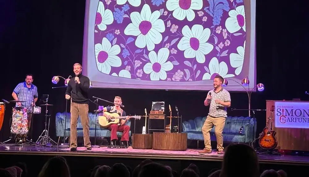 A band is performing on stage with floral patterns projected behind them and a sign reading The Sound of Simon  Garfunkel is visible