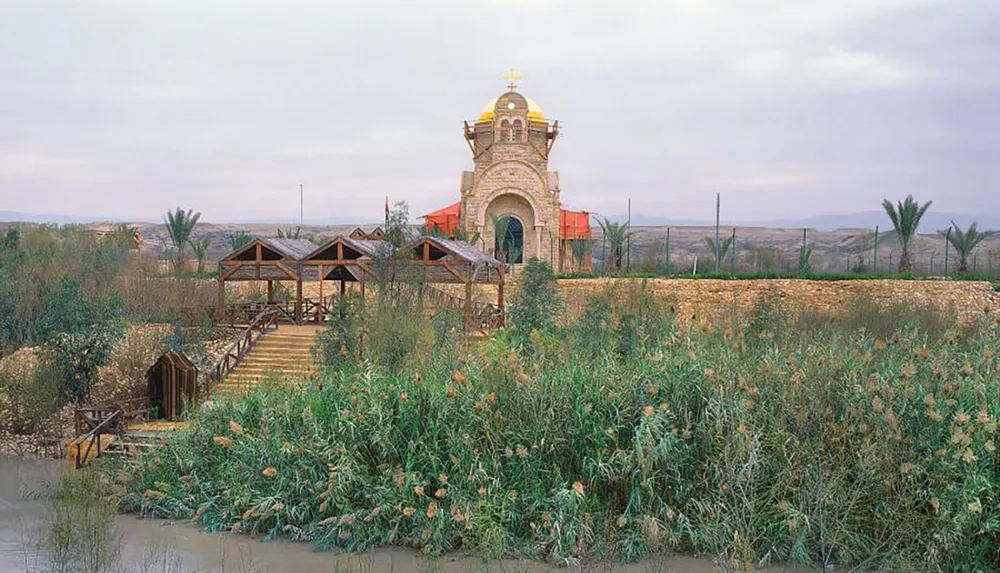 The image displays a small orthodox church with a golden dome surrounded by lush greenery and approached by a wooden bridge over a river