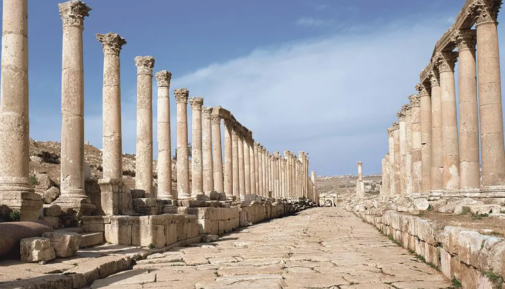 The image shows a long ancient colonnaded street lined with towering stone columns under a clear blue sky likely part of a historical or archaeological site
