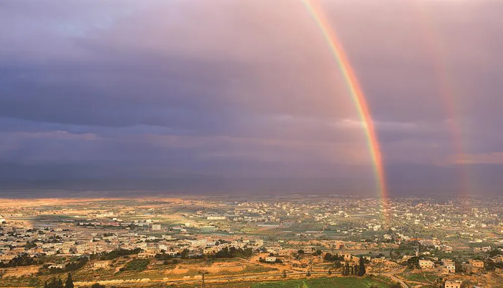 A vivid double rainbow arcs over a sprawling landscape dotted with buildings and fields under a dramatic sky