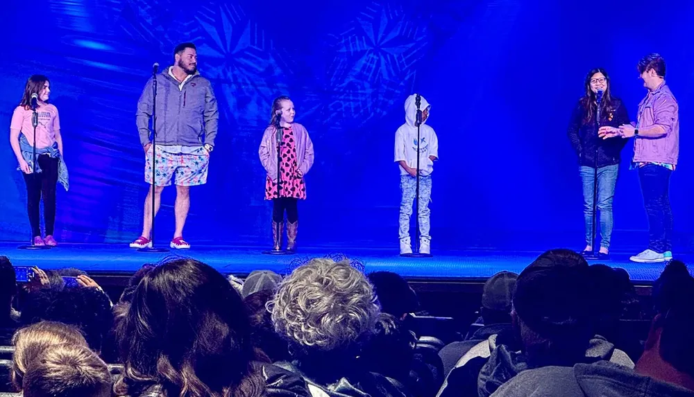 Several children are standing on stage possibly participating in an event or performance while an audience watches them from the darkened theater seats