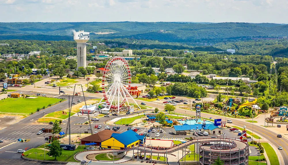 The image shows an aerial view of a vibrant amusement park featuring a large Ferris wheel roller coaster tracks and colorful buildings set against a backdrop of lush green hills