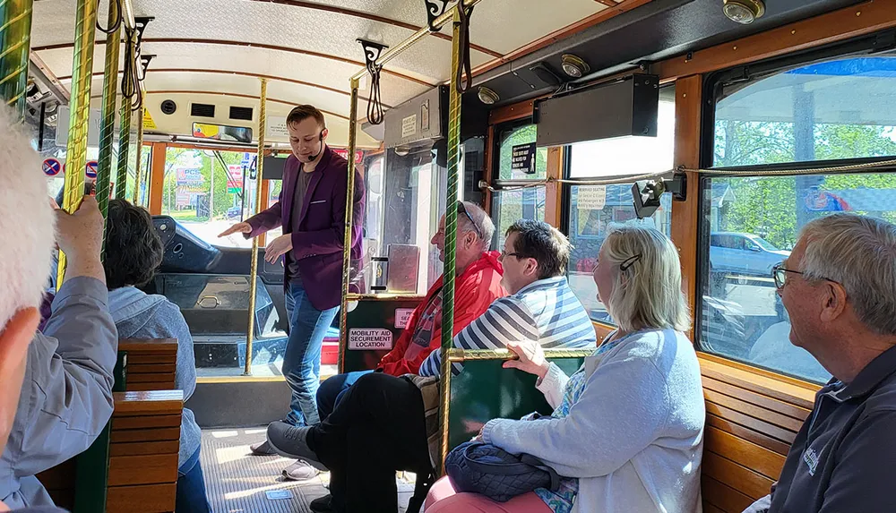 A group of passengers is attentively listening to a presentation from a standing individual inside a vintage-style trolley car