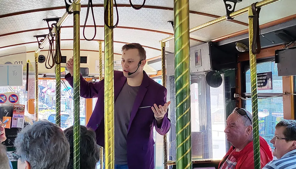 A person is giving a tour or presentation using a headset microphone to a group of seated passengers on a vintage-styled trolley or bus