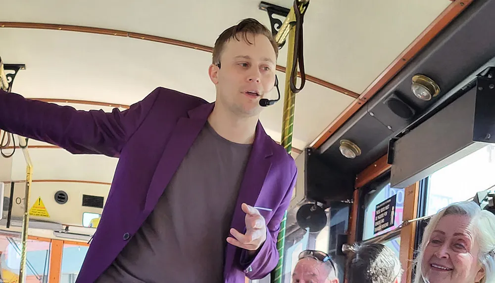 A man wearing a purple blazer and headset microphone is addressing an audience while holding onto a pole in what appears to be the interior of a bus or tram