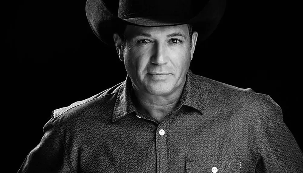 The image depicts a black and white portrait of a man wearing a cowboy hat and a patterned shirt gazing directly at the camera against a black background