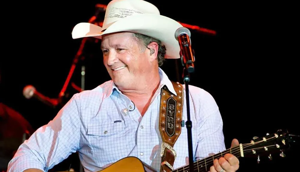 A smiling musician wearing a white cowboy hat is performing with an acoustic guitar on stage