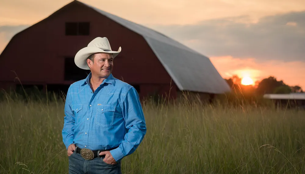 A man in a cowboy hat and blue shirt stands confidently in a field with a red barn and sunset in the background