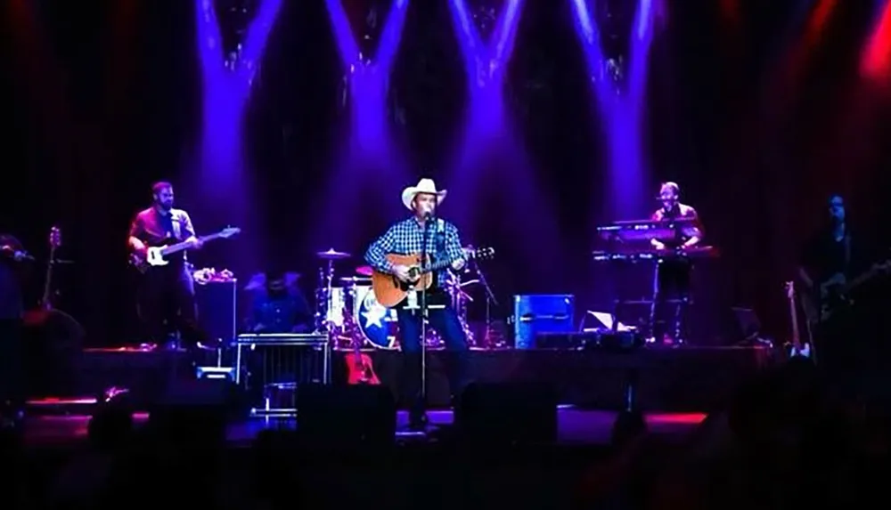 A band is performing on stage under blue and purple lighting with the central singer wearing a cowboy hat and playing an acoustic guitar