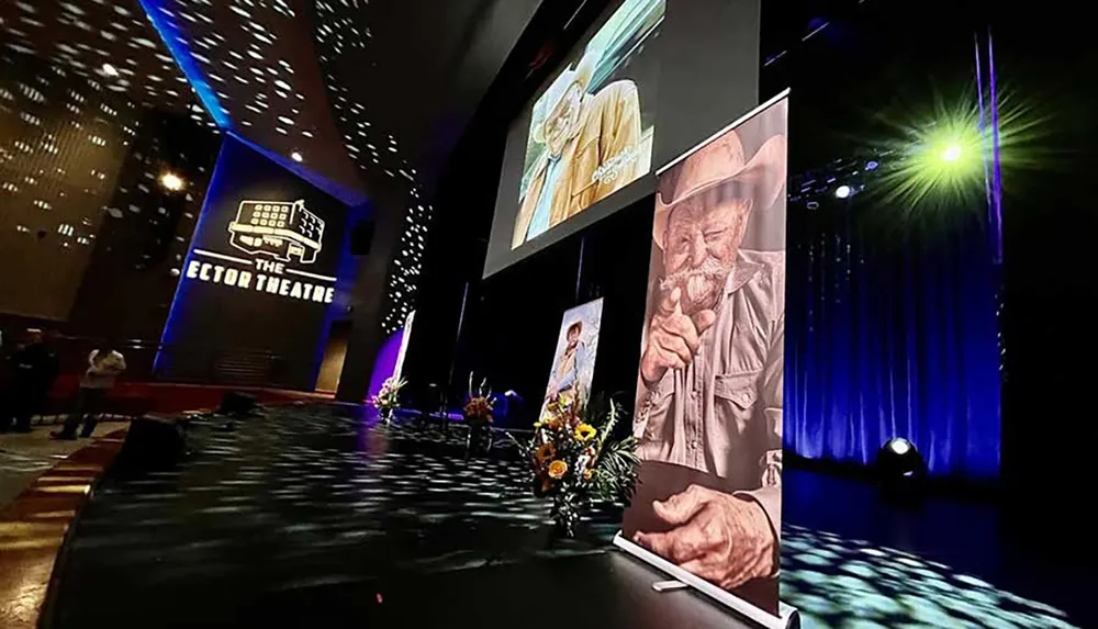 This image shows the interior of a theater with a large screen displaying a tribute image of an older individual likely during a memorial service or commemorative event