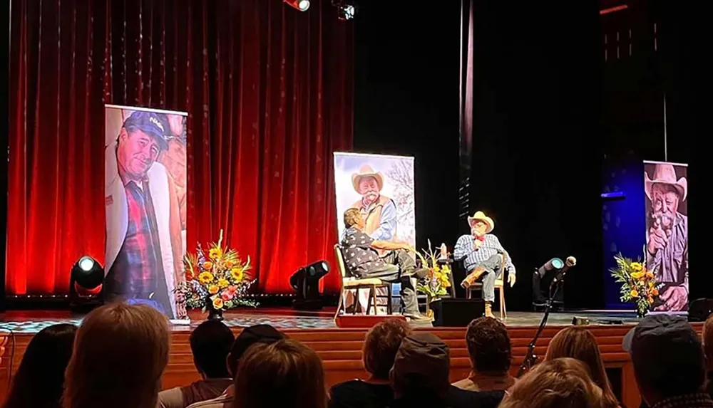 An individual in a cowboy hat is engaged in conversation with another person on stage during an event flanked by large portrait banners and watched by an audience