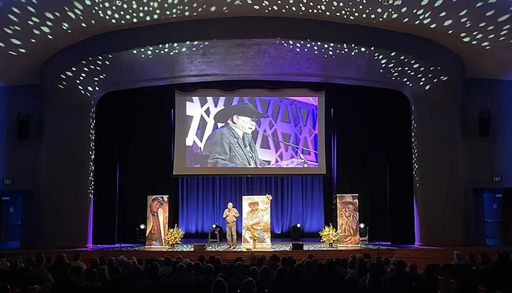 The image shows an event inside an auditorium with a speaker on stage an audience in attendance and a large screen displaying the speaker for better visibility