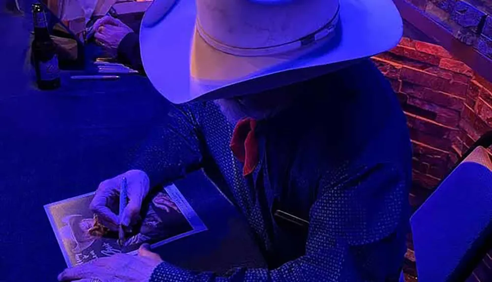 A person in a white cowboy hat and blue attire is autographing a photograph under blue lighting