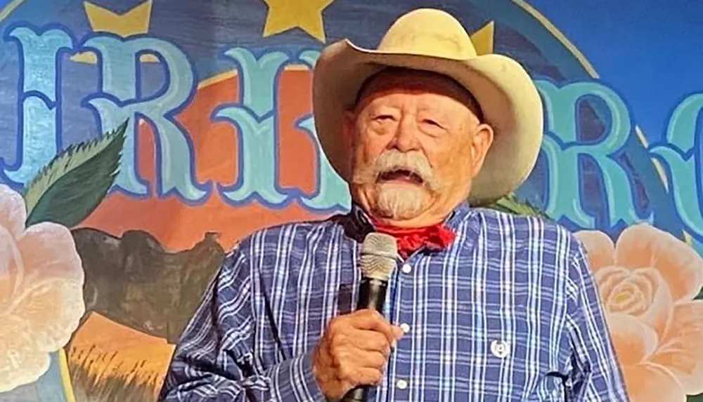 A person in a cowboy hat and bandana is speaking into a microphone against a colorful backdrop with the word Fiesta