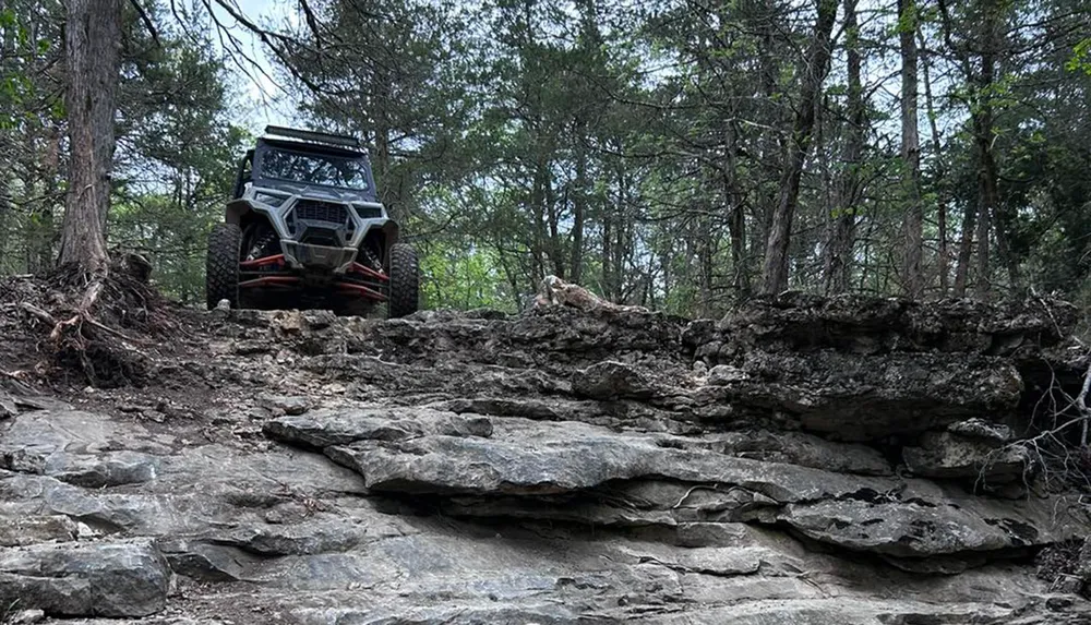 An off-road vehicle is positioned at the edge of a rocky ledge in a forested area