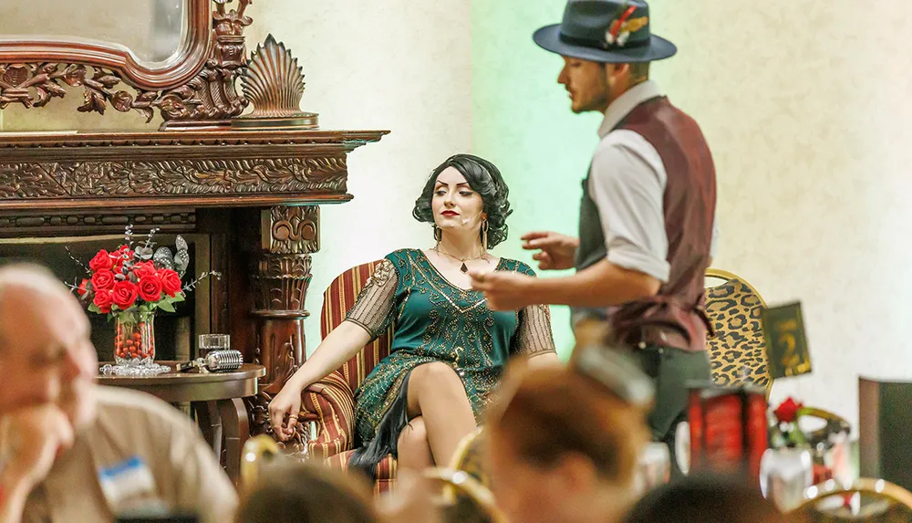 A woman in vintage attire sits elegantly in a chair listening to a man in a hat and vest in a room with a carved wooden fireplace and vibrant floral arrangements