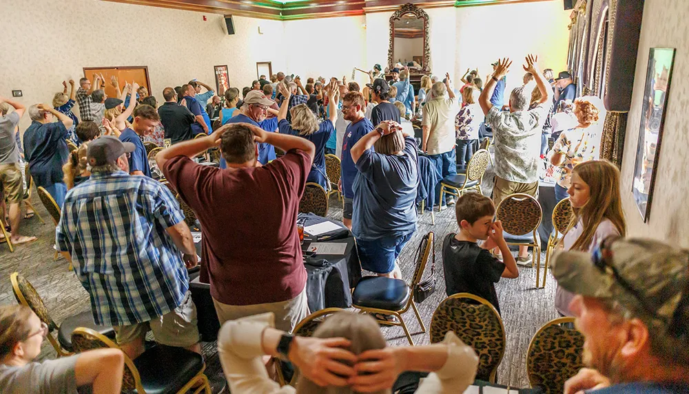 A large group of people is gathered indoors with many having their hands on their heads suggesting participation in an activity or game