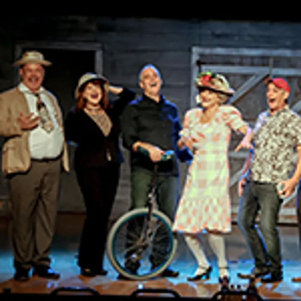 Five people are standing on stage in theatrical costumes with one person holding a unicycle
