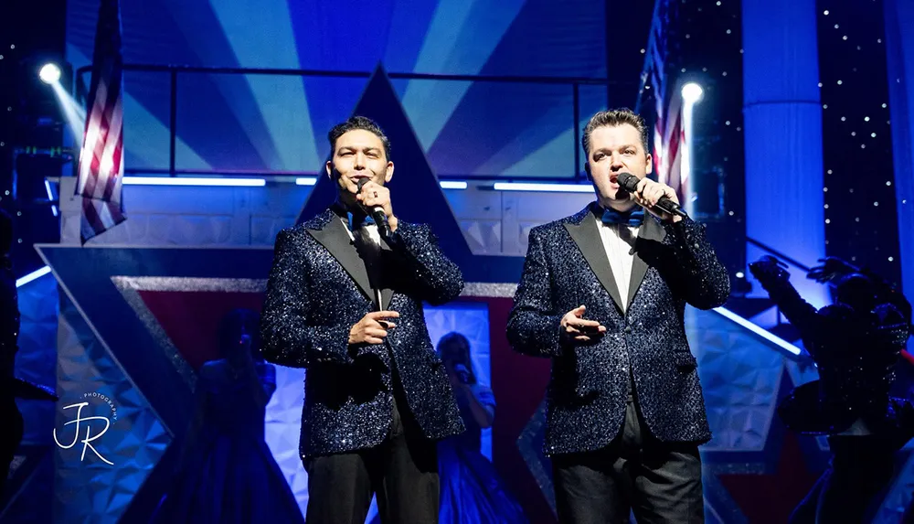 Two men in sparkling suits perform on stage with microphones against a backdrop featuring American flags and a geometric design