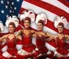 A group of dancers in red costumes pose in front of the American flag wearing white feathery headpieces