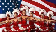 A group of dancers in red costumes pose in front of the American flag, wearing white feathery headpieces.