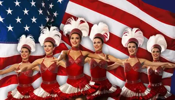A group of dancers in red costumes pose in front of the American flag, wearing white feathery headpieces.