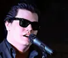 A person wearing sunglasses is singing into a microphone on stage