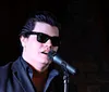 A person wearing sunglasses is singing into a microphone on stage
