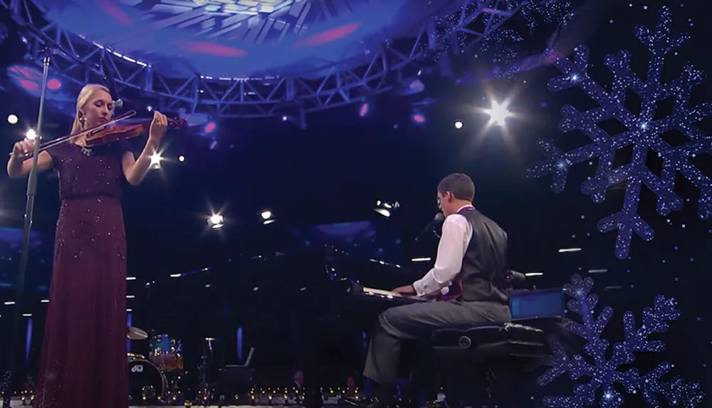 A woman in a maroon dress is playing the violin while a man in a vest is playing the piano set against a festive stage with snowflake decorations