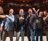 A group of six men poses and smiles on stage in front of an applauding audience in a theater