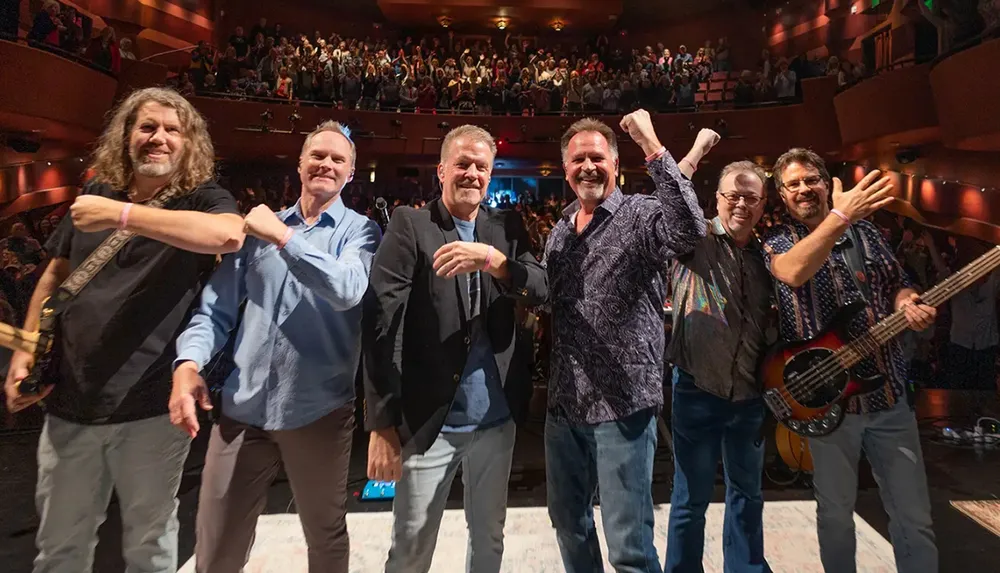 A group of six men poses and smiles on stage in front of an applauding audience in a theater
