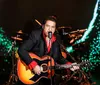 A musician is performing on stage with a guitar and microphone dressed in a black suit with red accents against a dynamic colorful background