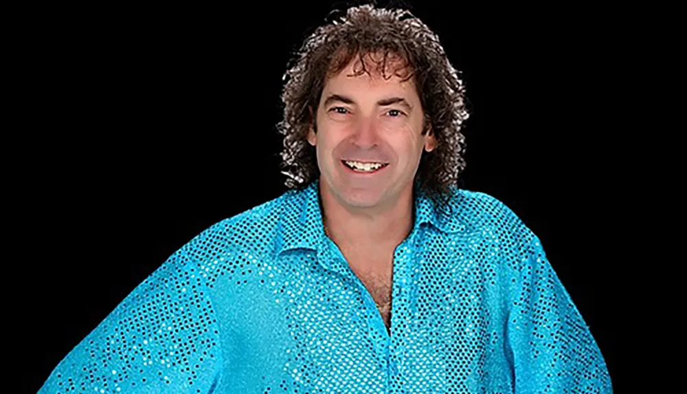 A person is smiling while wearing a sparkly blue shirt against a black background