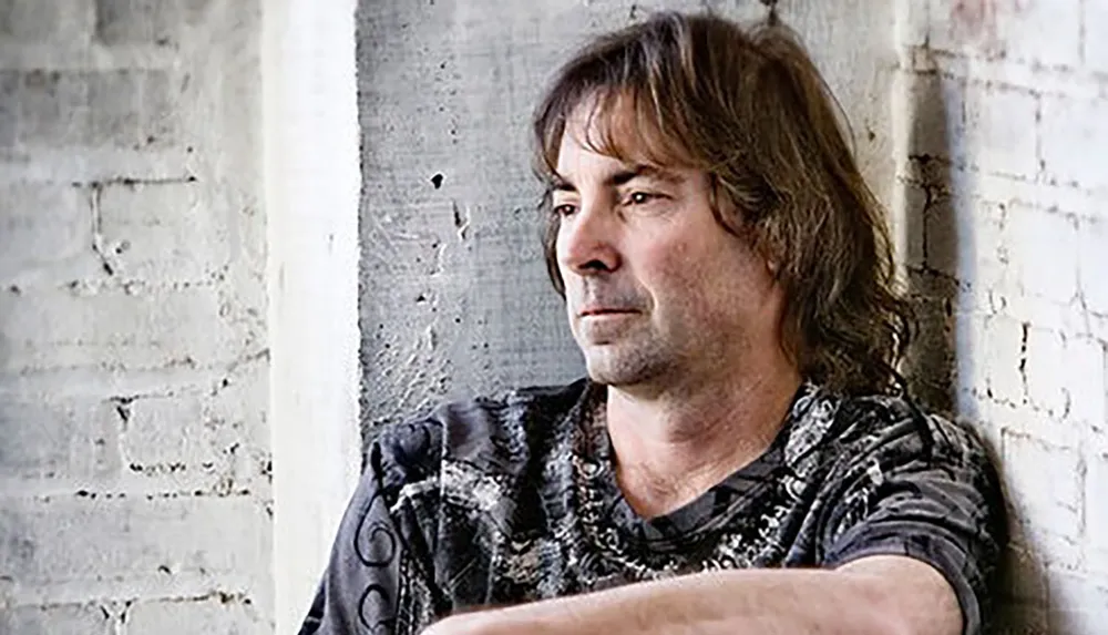 A man with shoulder-length hair wearing a patterned shirt is seated against a brick wall looking to the side