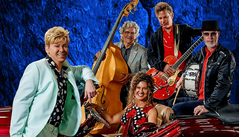A band of five people dressed in retro style pose with musical instruments against a blue background