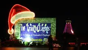 The image shows a festive entrance to 'The Trail of Lights' with a large, brightly-lit sign, a decorated structure that resembles a Santa hat, and a tower adorned with colorful lights in the background at night.