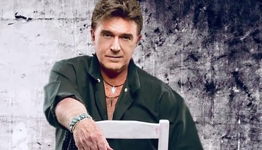 A man wearing a dark shirt and bracelets poses with his arm on the back of a chair against a textured background.