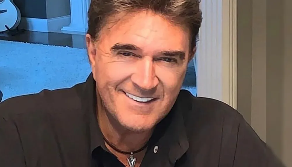 A man with short hair smiles warmly dressed in a dark shirt with a guitar visible in the background