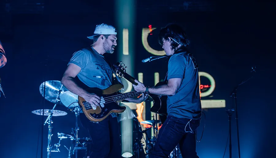 Two musicians are performing on stage, one playing a bass guitar and the other a guitar, with drum equipment visible in the background.