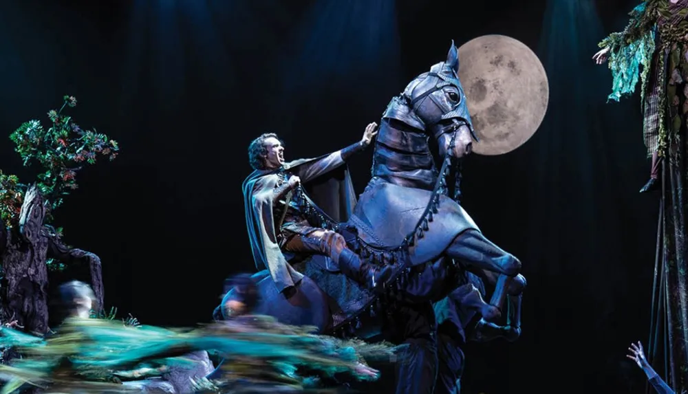 A person in theatrical attire rides a rearing horse puppet on stage set against a backdrop featuring a large full moon