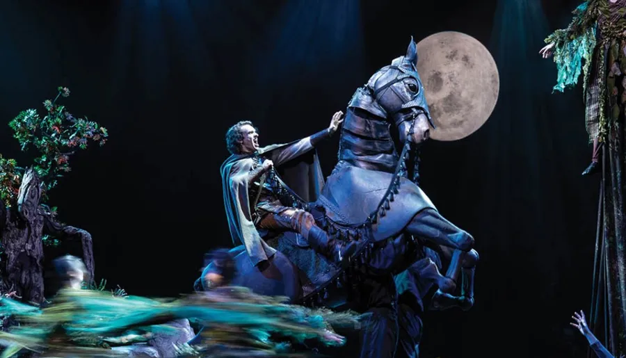 A person in theatrical attire rides a rearing horse puppet on stage, set against a backdrop featuring a large full moon.