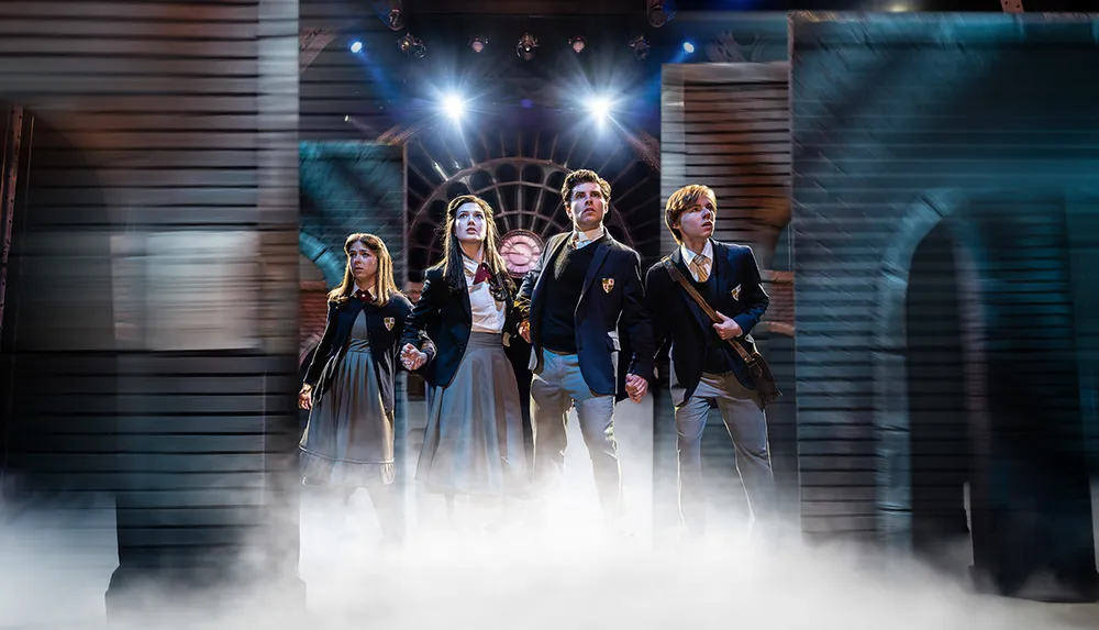 A group of four people in school uniforms stand together in a dramatic misty theater setting with bright lights behind them