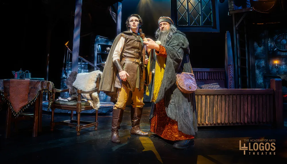 Two actors in medieval-style costumes perform on a stage with rustic set pieces and lighting