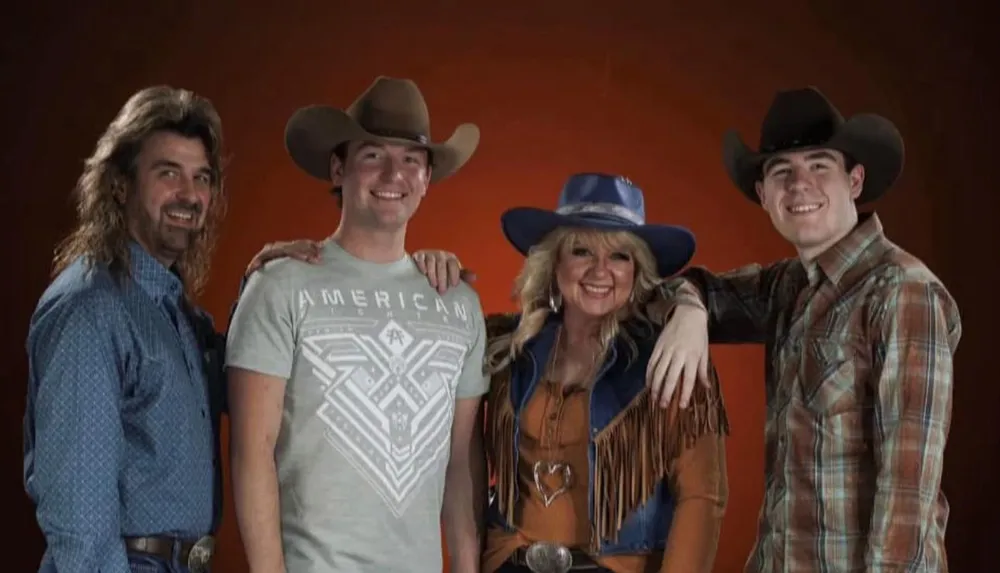Four people dressed in Western attire with cowboy hats smile for the camera against a warm-toned backdrop