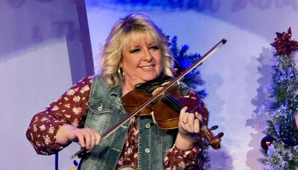 A woman with blonde hair is smiling while playing the violin wearing a denim vest over a floral shirt with Christmas decorations in the background