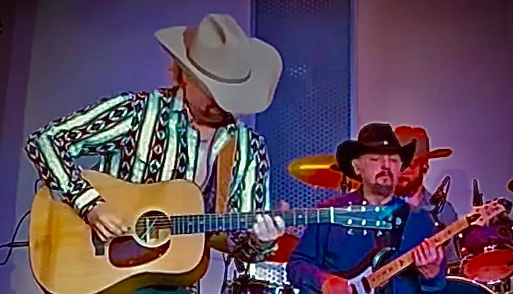 Two musicians wearing cowboy hats perform on stage one playing an acoustic guitar and the other an electric guitar