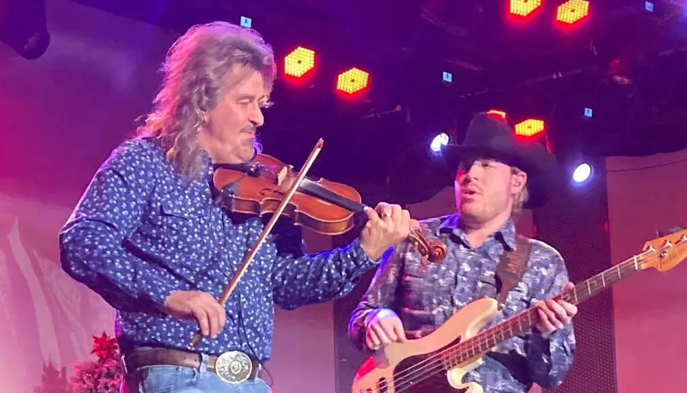 Two musicians are performing on stage one playing a violin and the other playing a bass guitar both wearing patterned shirts and illuminated by colorful lights