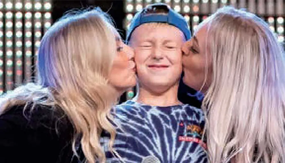 A young person in a blue tie-dye shirt and backwards cap is being kissed on both cheeks by two women