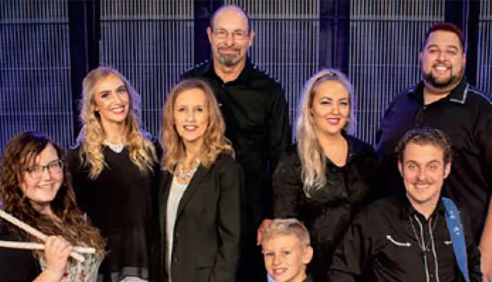 Seven adults and one child are posing together smiling with one person holding drumsticks