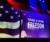 A person stands on stage beside a large screen displaying the American flag and the words Cherish Defend Freedom